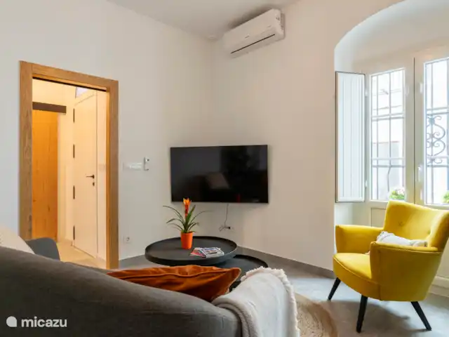 Ruime woonkamer met 2-persoonsbank, fateuil, werktafel met hoge stoel, TV en airco/verwarming.